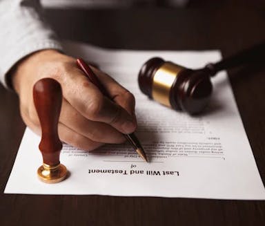 A hand writes a last will and testament. On the will is a gavel and a stamp.