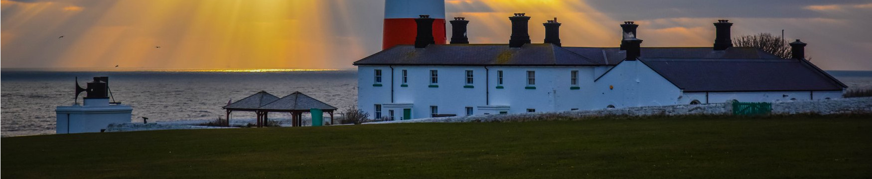 Dawn breaks behind the base of a lighthouse and a row of houses. Photo by Trevor Liddell